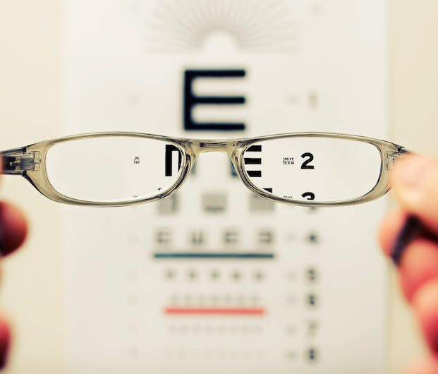 Es wird das Sehfeld einer Person mit Sehschwäche abgebildet. Man sieht eine Brille und im Hintergrund sehr unscharf einen Sehtest. Nur durch die Brillengläser lässt sich scharf sehen.