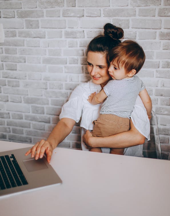 Frau mit Kleinkind auf dem Arm, bedient einen Laptop mit der rechten Hand.