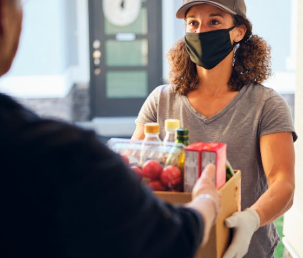 Junge Frau mit Maske überreicht Karton mit Lebensmitteln an der Haustür